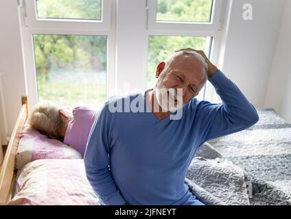 Uomo anziano seduto a letto e stretching al collo al mattino mentre la donna dorme ancora sullo sfondo Foto Stock