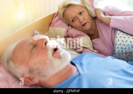 Uomo anziano che dorme sulla schiena a letto e che russava mentre la donna è sveglio e infastidito tenendo il cuscino sulle orecchie Foto Stock