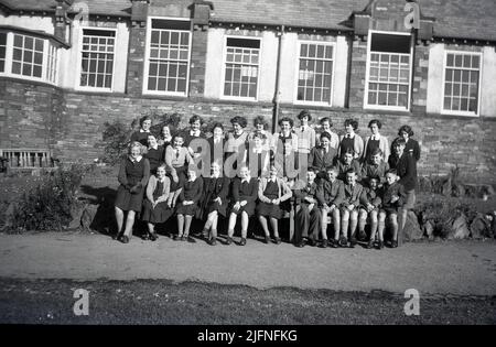 1951, storico, una grande classe di bambini eccitati nella loro uniforme, seduti e in piedi insieme fuori dal loro edificio scolastico per una foto di classe di gruppo, Inghilterra, Regno Unito. Foto Stock