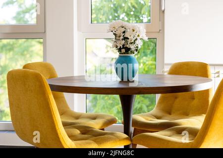 Vaso blu con fiori margaret su tavolo da pranzo rotondo in legno con sedie gialle accoglienti di fronte alle finestre a casa, arredamento moderno Foto Stock