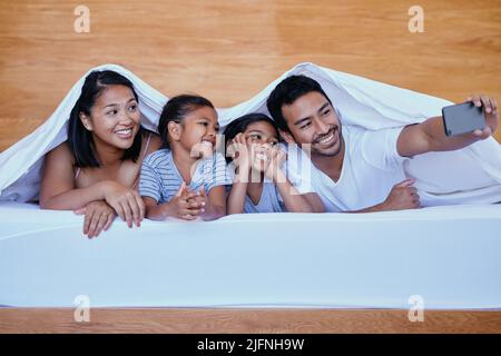 Felice famiglia a letto. Una giovane famiglia ispanica a letto con un selfie utilizzando uno smartphone. Due genitori che si legano con le loro figlie a casa. Sorelle Foto Stock