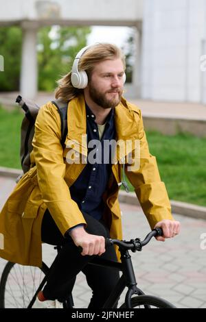 Giovane uomo bearded che ascolta la musica in cuffie wireless mentre si guida una bicicletta lungo la strada Foto Stock