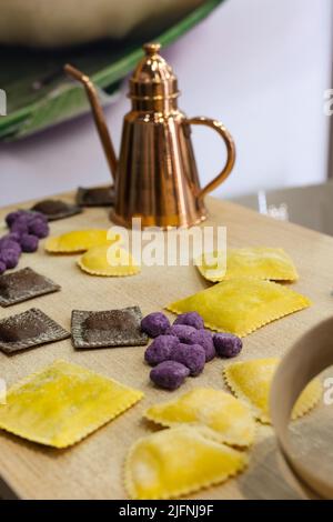 Tortelli crudi appena fatti e altri tipi di pasta italiana su tavola di legno e una caffettiera in rame sullo sfondo. Foto Stock