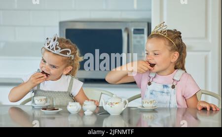 Due bambine che hanno una festa del tè della principessa a casa. Amici fratelli sorelle che indossano le tiaras mentre giocano con il set di tè e hanno biscotti in cucina Foto Stock