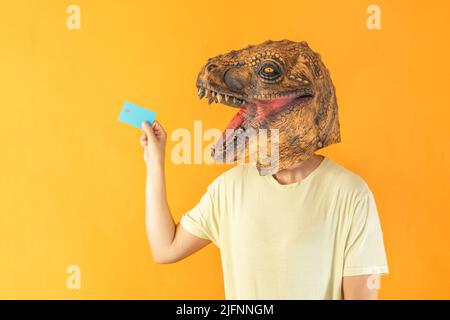 Ritratto di giovane donna in dinosauro animale testa maschera tenendo carta di credito su sfondo arancione Foto Stock