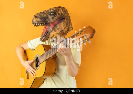 Donna irriconoscibile che suona la chitarra indossando maschera animale dinosauro isolato su sfondo arancione studio Foto Stock