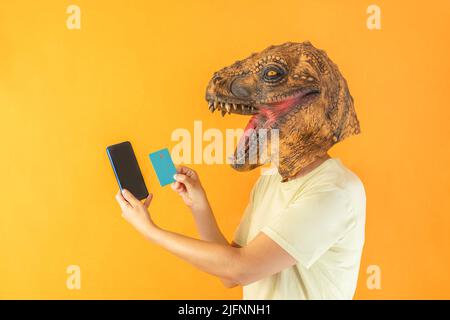Felice persona irriconoscibile indossando maschera testa animale dinosauro, tenendo smartphone e carta di credito su sfondo arancione Foto Stock