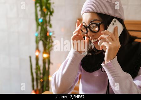 Giovane donna asiatica in occhiali ridendo e parlando al telefono cellulare al caffè Foto Stock