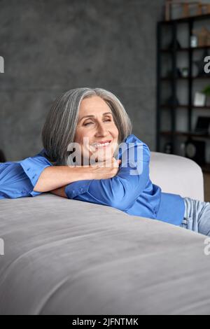 Sorridente vecchia donna matura e riposante sognando seduta sul divano a casa. Foto Stock