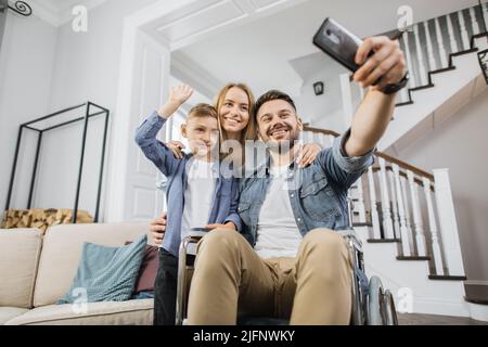 Famiglia sorridente in piedi vicino al padre con esigenze speciali e guardando la fotocamera dello smartphone durante le videochiamate online. Figlio in età scolastica che agitava la mano mentre il papà in sedia a rotelle tiene il telefono. Foto Stock