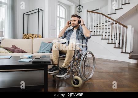 Il giovane usa le cuffie mentre si siede in sedia a rotelle nel luminoso soggiorno e ascolta nuove canzoni. Maschio bearded sensazione di calma durante il relax con la musica a casa. Foto Stock