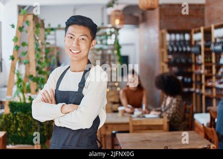 Ritratto di un sorridente bel proprietario di affari asiatici in piedi con le braccia incrociate nel suo caffè. Uomo di gara misto sicuro e di successo indossando un Foto Stock