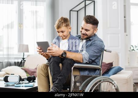 Papà disabile seduto in sedia a rotelle e che tiene il figlio in ginocchio mentre gioca su un tablet moderno in un luminoso soggiorno. Famiglia caucasica amichevole giocare insieme a casa. Foto Stock