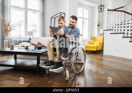 Sorridendo papà con esigenze speciali seduto in sedia a rotelle e tenendo il figlio in ginocchio mentre gioca sul tablet digitale. Famiglia positiva che utilizza un dispositivo portatile per giocare a giochi educativi per bambini. Foto Stock