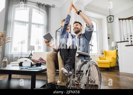 Ragazzo della scuola che dà i cinque alti al papà disabile dopo la vittoria sull'app del tablet mentre si siede sulle ginocchia dei padri. Concetto di supporto familiare e tecnologia moderna. Foto Stock