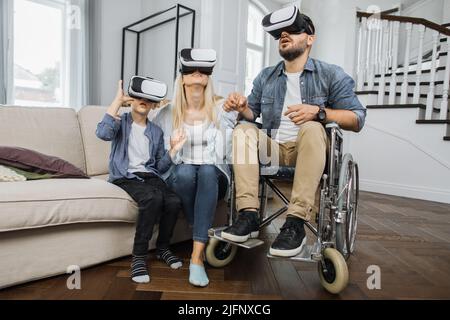 Una famiglia felice che utilizza la moderna tecnologia futuristica in un luminoso soggiorno. Padre caucasico in sedia a rotelle che insegna a giovane moglie e figlio carino come utilizzare la cuffia per realtà virtuale. Foto Stock