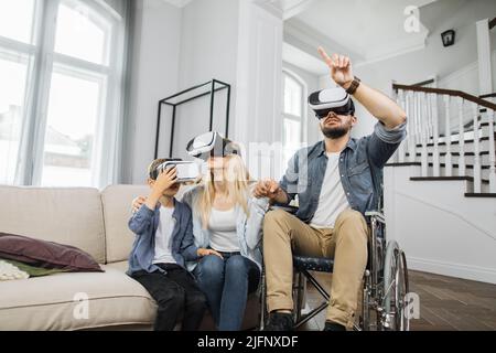 Una famiglia felice che utilizza la moderna tecnologia futuristica in un luminoso soggiorno. Padre caucasico in sedia a rotelle che insegna a giovane moglie e figlio carino come utilizzare la cuffia per realtà virtuale. Foto Stock