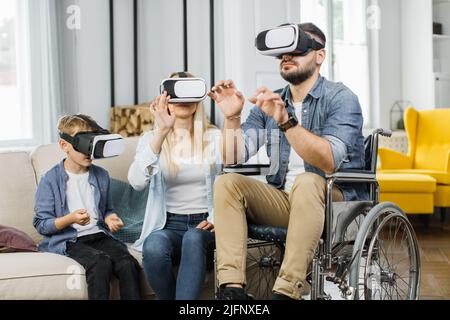 Famiglia amichevole di madre, figlio e padre in sedia a rotelle indossando cuffie VR nel luminoso soggiorno. Concetto di persone, disabilità e tecnologia futuristica. Foto Stock