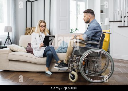 Medico caucasico di discussione medico rapporto con l'uomo in sedia a rotelle in soggiorno. Fisioterapista competente che prende appunti sulla clipboard mentre parla con il paziente di sesso maschile. Foto Stock