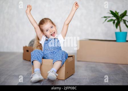 Una bambina bionda gioca a casa in quarantena con scatole di cartone. Il bambino si sposta in un nuovo appartamento, è felice e felice. Foto Stock