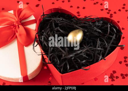 Uovo d'oro in una confezione regalo a forma di cuore su sfondo rosso con paillettes d'oro. Capodanno cinese , esclusività, scelta migliore, investimenti, Banca. Foto Stock