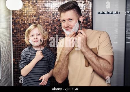 Ritratto di felice padre che radera con il ragazzino che imita il papà e sorride Foto Stock