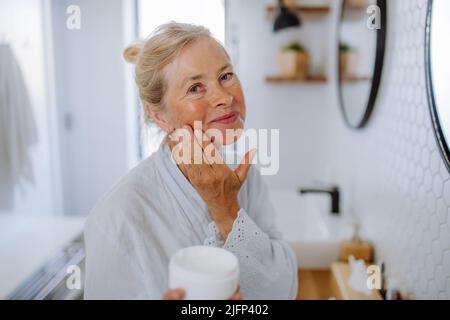 Bella donna anziana in accappatoio applicazione crema naturale in bagno, cura della pelle e mattina concetto di routine. Foto Stock