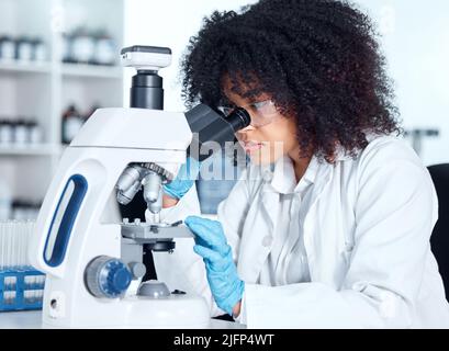 Giovane donna afroamericana che indossa un camice da laboratorio e occhiali che guarda campioni medici su un microscopio nel suo laboratorio. Una scienziata femminile mista Foto Stock