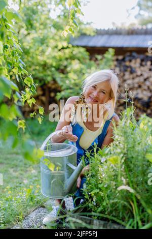 Donna anziana felice che prende la cura dei fiori all'aperto in giardino, innaffiare con la lattina. Foto Stock