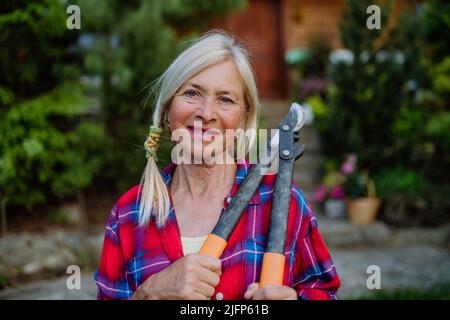 Ritratto di donna anziana giardinaggio in estate, tenendo potare sheares e guardando la macchina fotografica. Foto Stock