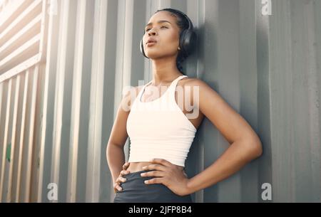Una ragazza ispanica vestita con cuffie e una pausa di riposo per prendere il respiro dopo una corsa o jogging in un ambiente urbano all'aperto. Femmina Foto Stock