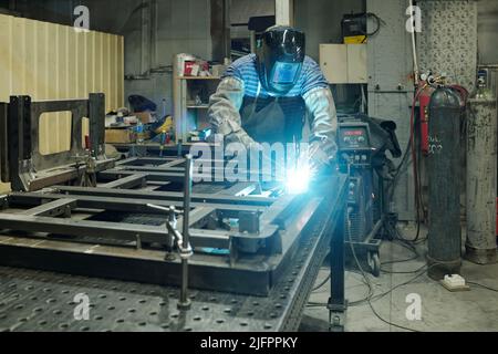 Giovane specialista maschile in abbigliamento da lavoro di protezione saldatura di parti metalliche di enorme macchina industriale mentre si piega sul suo banco di lavoro Foto Stock