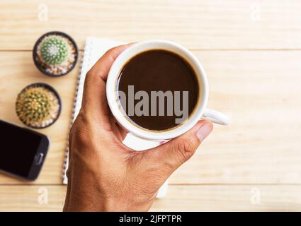 Mano che raggiunge per tazza di caffè e smartphone sfocato, nota libro su sfondo tavolo in legno. Vista dall'alto. Foto Stock
