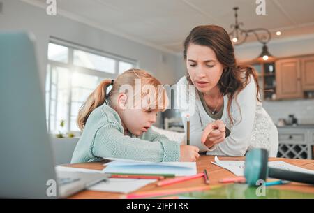 Adorabile ragazza caucasica seduta a tavola e fare i compiti mentre sua madre la aiuta. Bella giovane donna seria che la indica e la insegna Foto Stock