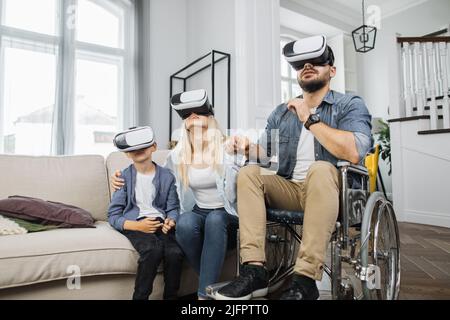 Famiglia amichevole di madre, figlio e padre in sedia a rotelle indossando cuffie VR nel luminoso soggiorno. Concetto di persone, disabilità e tecnologia futuristica. Foto Stock