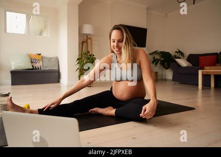 Incinta bianca caucasica giovane femmina sorridente, seguendo un tutorial online di stretching mentre si siede su tappeto yoga in soggiorno moderno Foto Stock