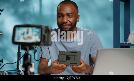 Influencer che esamina la batteria di alimentazione con montaggio a V sulla telecamera per consigli sul canale. Vlogger filma video per rivedere il caricatore portatile ricaricabile v lock, utilizzando treppiede e attrezzatura. Foto Stock