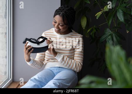 Giovane donna nera eccitata seduta sul davanzale guardando curiosamente il visore VR nelle mani Foto Stock
