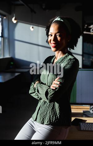 Ritratto di sorridente giovane donna d'affari afroamericana seduta con le braccia incrociate in ufficio Foto Stock