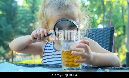 Odessa, Ucraina, Europa dell'Est. 4th luglio 2022. La ragazza piccola carina guarda con attenzione nell'obiettivo al succo nel bicchiere. Primo piano della ragazza bionda che studia il suo succo in vetro mentre la guarda attraverso la lente d'ingrandimento mentre si siede in strada caffè sul parco. (Credit Image: © Andrey Nekrasov/ZUMA Press Wire) Foto Stock