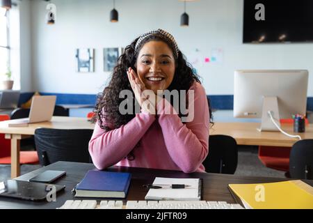 Ritratto di sorridente biraciale giovane donna d'affari seduta alla scrivania in ufficio creativo Foto Stock