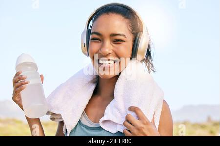 Donna eccitata sorridendo e ascoltando musica mentre fuori per un allenamento nella natura. Felice giovane atleta femminile con cuffie wireless, bottiglia d'acqua e. Foto Stock