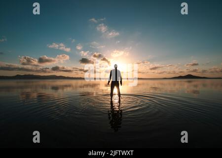 Silhouette di un uomo in piedi con la schiena rivolta al tramonto sulle acque poco profonde del Mar Menor, regione di Murcia, Spagna, creando increspature sul Foto Stock