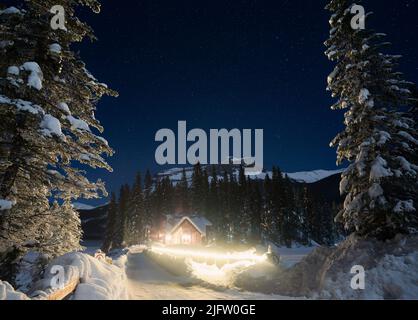 Piccola capanna in una foresta d'inverno circondata da alberi, montagne con stelle luminose sopra, Yoho NP, Canada Foto Stock