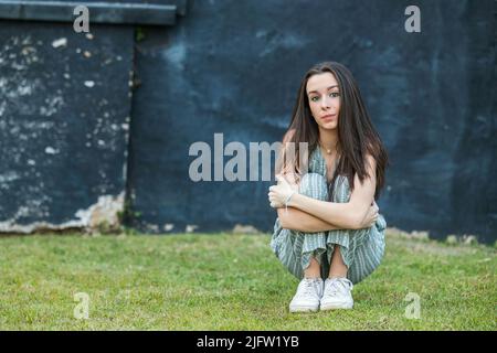 Una bella ragazza bruna teen con uno sguardo serio seduta e appoggiata contro un muro in pensiero con un blu nero ruvido muro della città dietro di lei Foto Stock