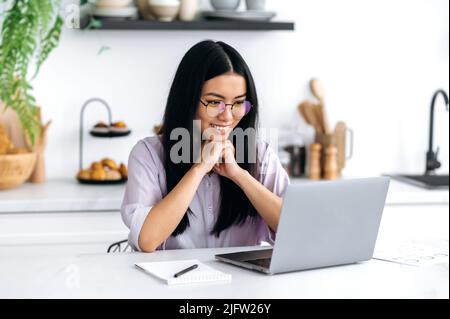 Bella ragazza felice di nazionalità asiatica, una donna cinese freelance, uno studente con occhiali, si siede a un tavolo in cucina, guarda lo schermo del computer portatile, sorride, si rallegra per le buone notizie, un buon affare Foto Stock