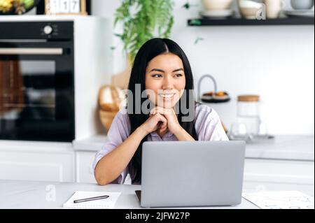 Bella bella ragazza giovane bruna carina di nazionalità asiatica, in eleganti abiti casual, freelancer, designer, si siede a un tavolo con un computer portatile in cucina a casa, guarda via, sorride amichevole Foto Stock