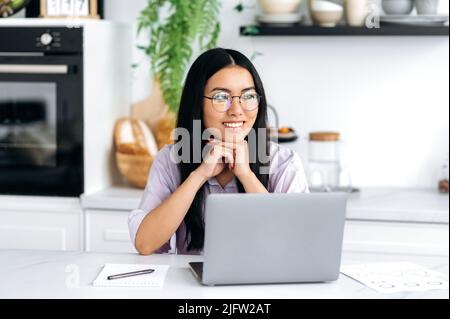 Bella bruna smart cinese giovane donna con occhiali, in elegante casual abbigliamento, freelancer, designer, content manager, si siede a un tavolo con un portatile in cucina a casa, si guarda via, sorride amichevole Foto Stock