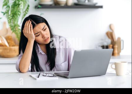 Stanco esausta ragazza cinese triste, freelancer, designer creativo, content manager, sembra deluso per lo schermo del laptop, appoggiata la testa sulla sua mano, non ha il tempo di completare il progetto in tempo Foto Stock