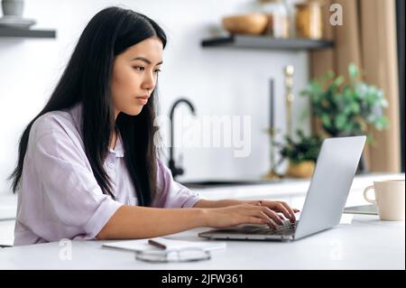 Giovane donna cinese intelligente pensieroso, sedersi a casa in cucina, lavorare o studiare online, ha messo a fuoco lo schermo del laptop, progettazione e analisi progetto futuro, startup, studentessa femminile che fa i compiti Foto Stock